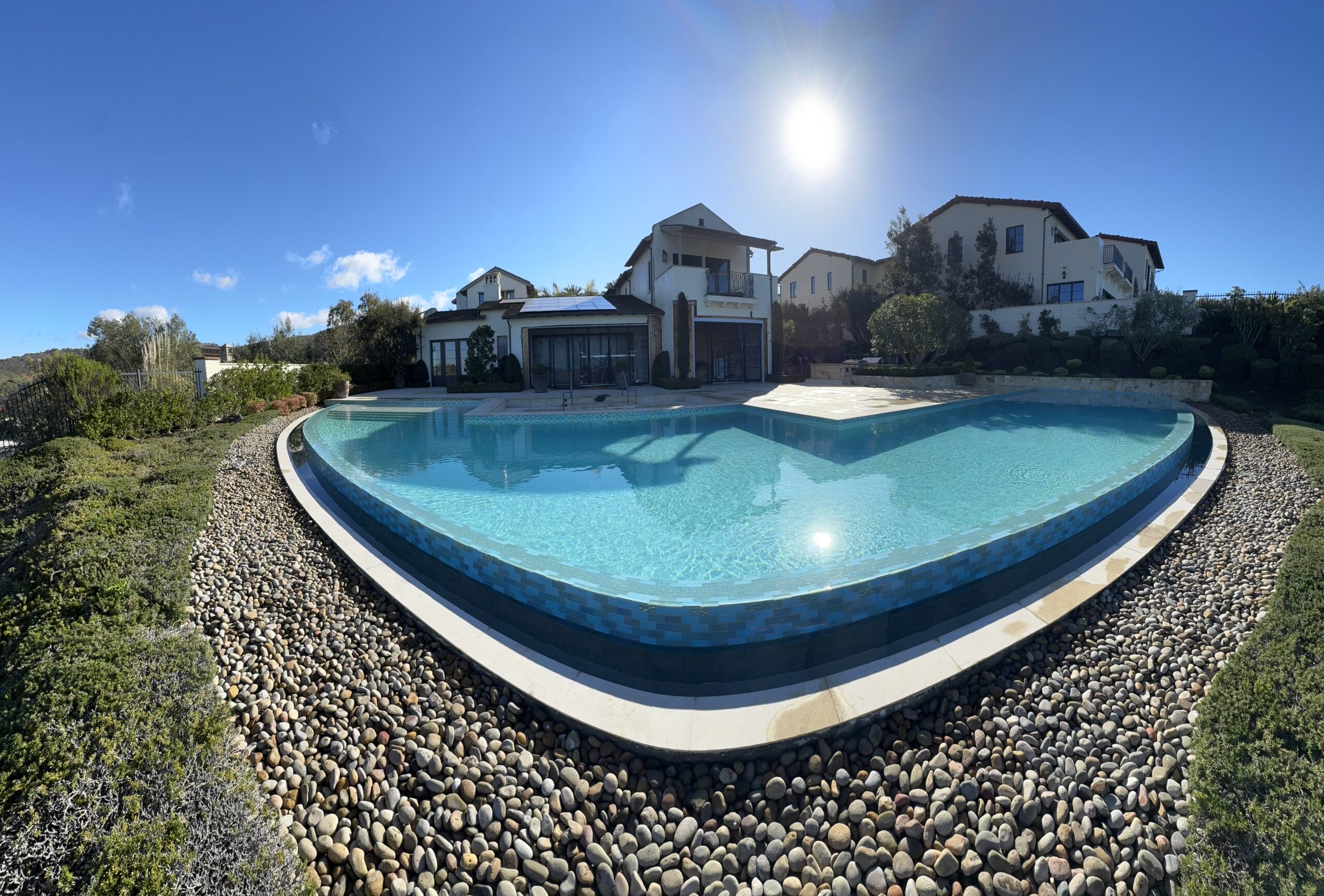 Large outdoor swimming pool with clear water, surrounded by rocks and greenery, set in front of Mediterranean-style houses under a bright sunny sky.